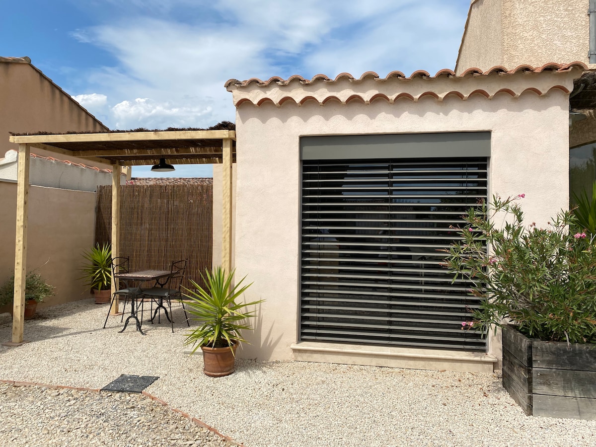 An independent studio is shown, featuring a smooth stucco exterior and large horizontal blinds. A seating area is set up under a wooden pergola, surrounded by decorative plants. Gravel pathways create a welcoming atmosphere, enhancing the connection to the outdoor space.