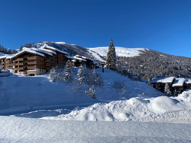 A snow-covered landscape provides a view of the resort buildings nestled in the hillside. Tall evergreen trees are visible, their branches laden with snow, under a clear blue sky. Rolling slopes in the background create a serene winter backdrop.