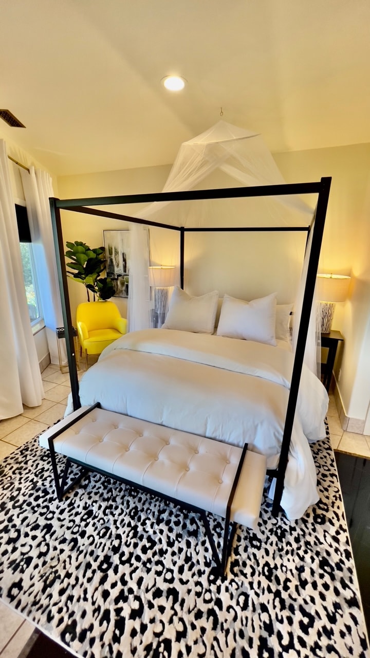 A modern bedroom features a spacious canopy bed adorned with white linens and decorative pillows. A stylish bench sits at the foot of the bed, placed on a leopard-print rug. Two bedside lamps provide soft illumination alongside large windows, allowing natural light to enter the room.