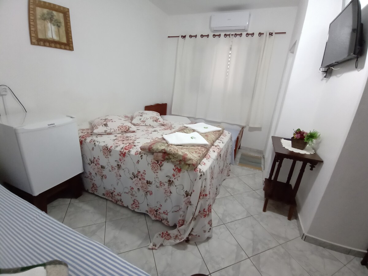 A cozy room features a floral-patterned bed covering over a spacious bed. A small fridge is positioned in the corner, while a wooden side table stands beside the bed. Light enters through sheer curtains, and a wall-mounted television is visible.