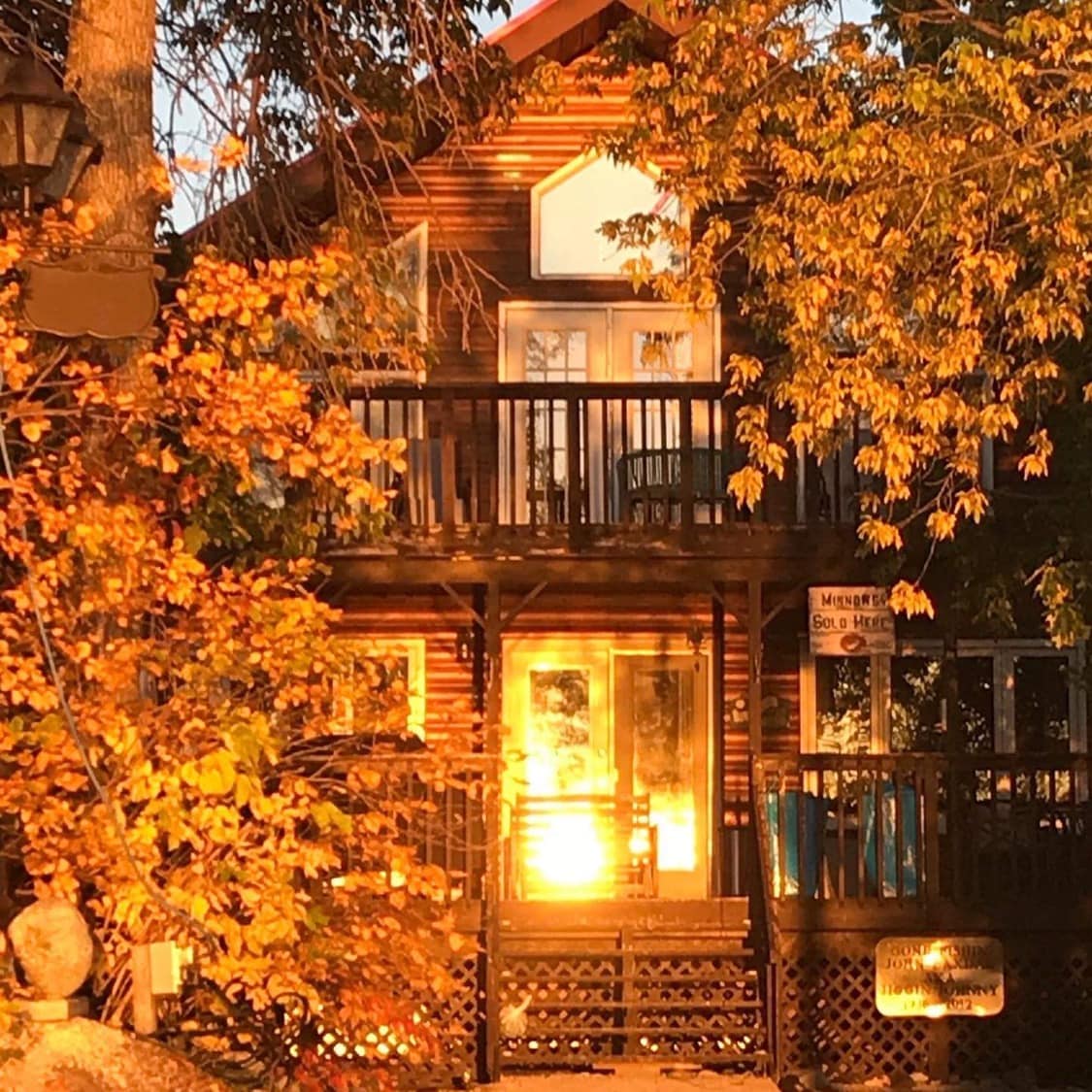 The rustic cottage is framed by autumn foliage, with warm light reflecting off large windows. A welcoming covered porch leads to a spacious deck, adorned with seating options. The wooden exterior complements the natural surroundings, creating a harmonious and inviting atmosphere.