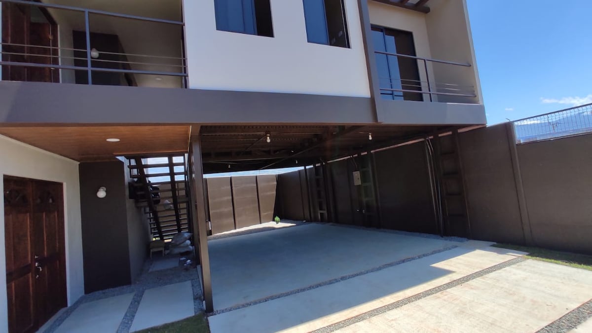 A modern structure features two levels, with a covered parking area below. The exterior is adorned with smooth walls and large windows, allowing ample natural light. Stairs lead to the upper level, with a spacious pathway and green grass surrounding the building.