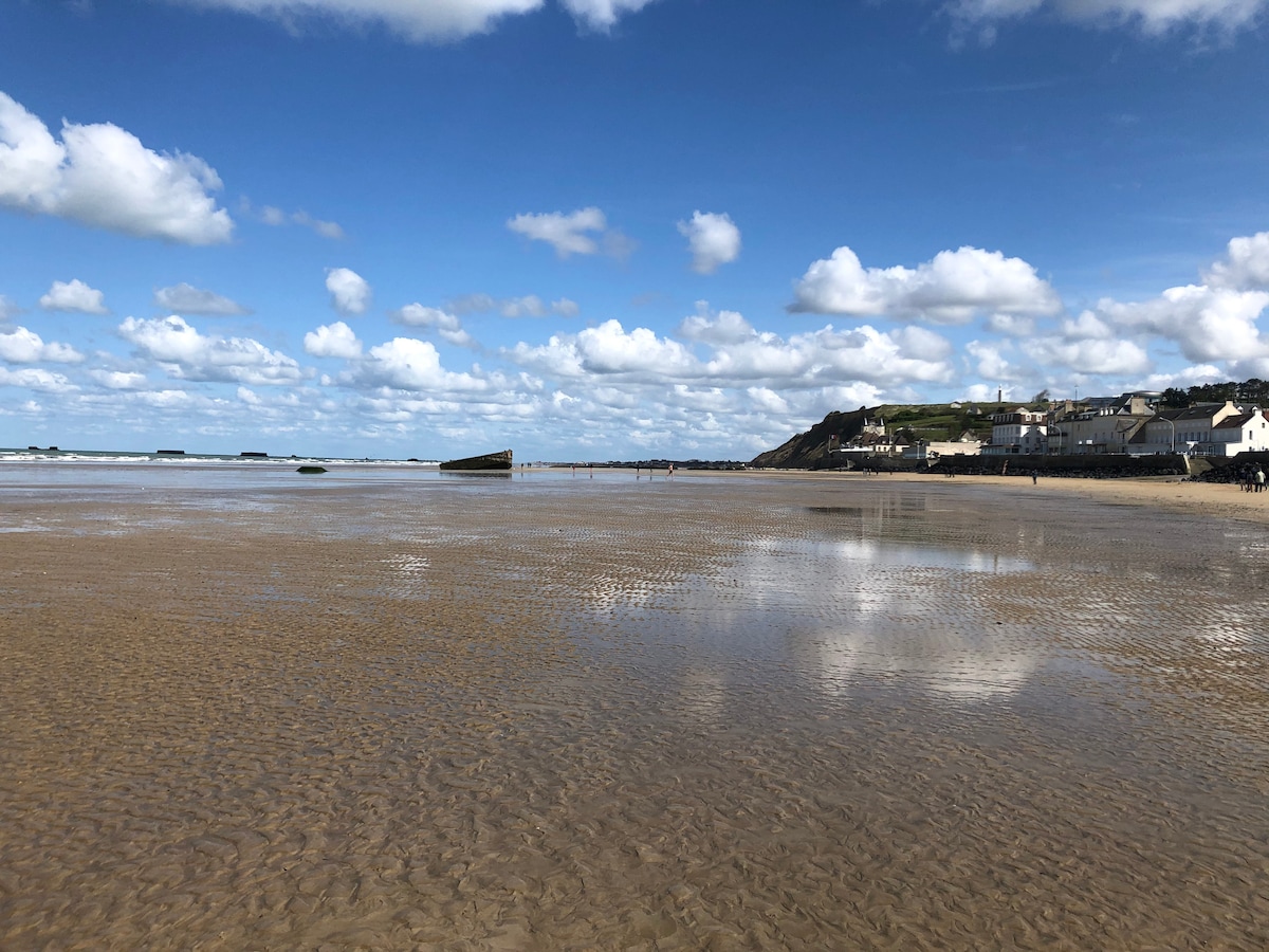 A vast sandy beach is displayed under a clear sky, characterized by scattered clouds. The tide has receded, revealing a patterned wet sand surface. In the distance, remnants of a shipwreck are visible, along with a coastal town featuring several buildings along the shore.