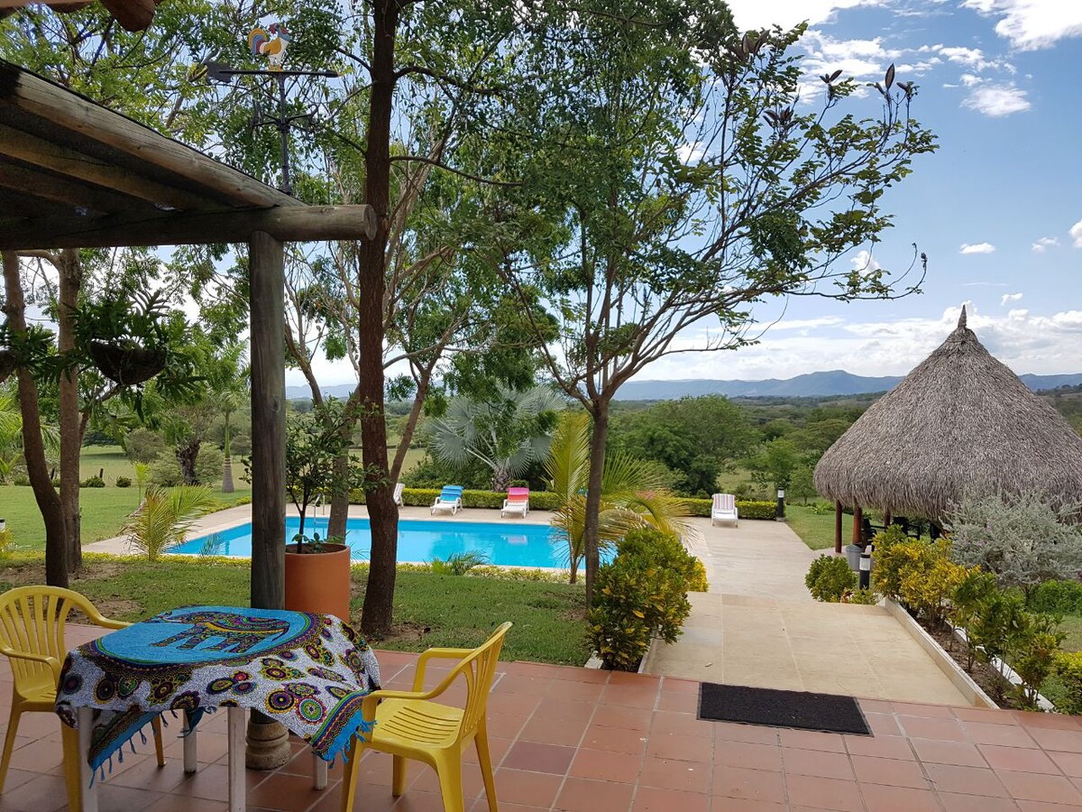 A shaded patio area features a round table adorned with vibrant patterns, complemented by a pair of yellow chairs. In the background, a clear pool is framed by lush greenery and a thatched gazebo, offering a serene outdoor setting.