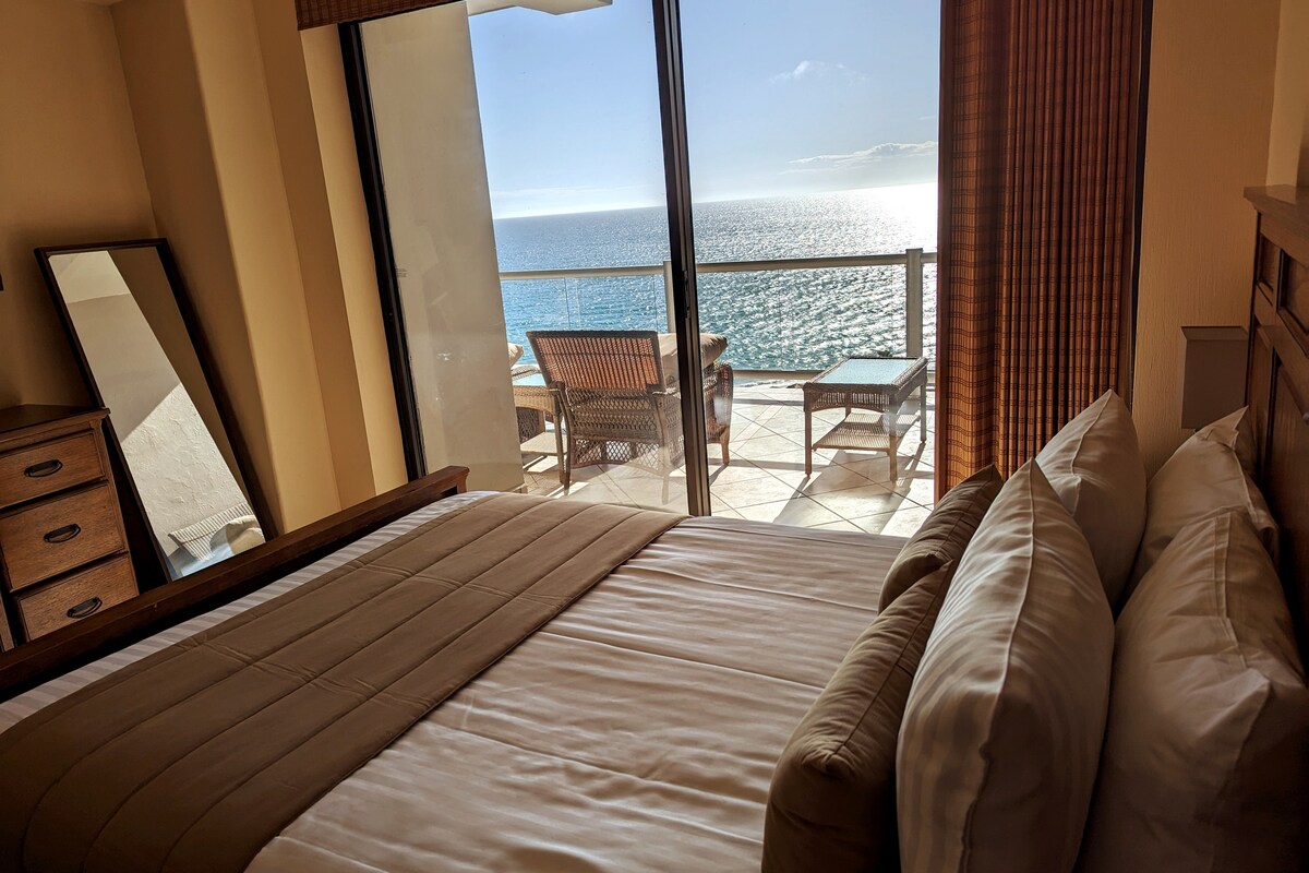 A spacious bedroom features a large bed with neutral linens, positioned to overlook the ocean through a floor-to-ceiling sliding glass door. A balcony with outdoor seating is visible, offering a serene view of the water and horizon beyond.
