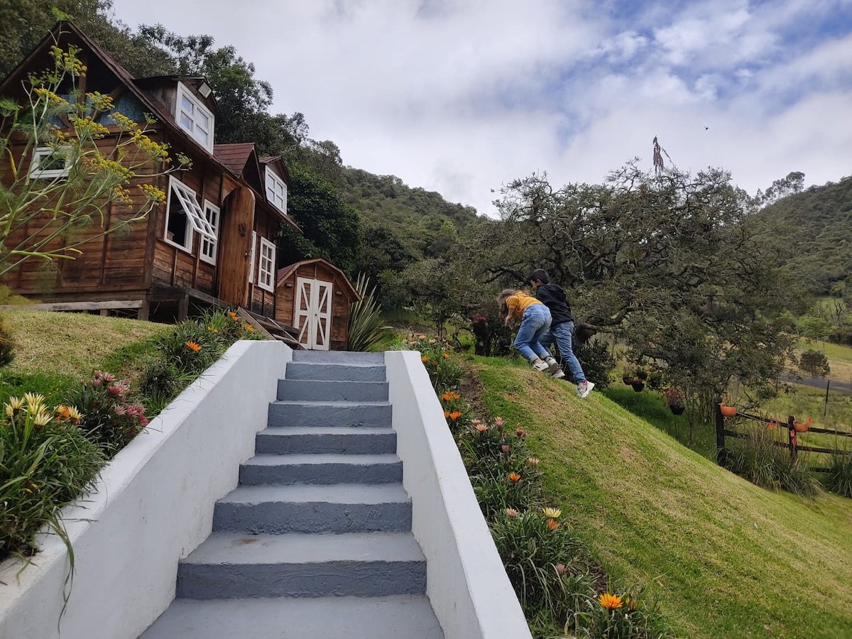 A set of wooden cabins is nestled among greenery and hills. Stone steps lead up to the cabins, with a small garden filled with colorful flowers on either side. Two individuals are seen exploring the hillside, surrounded by a serene natural landscape.