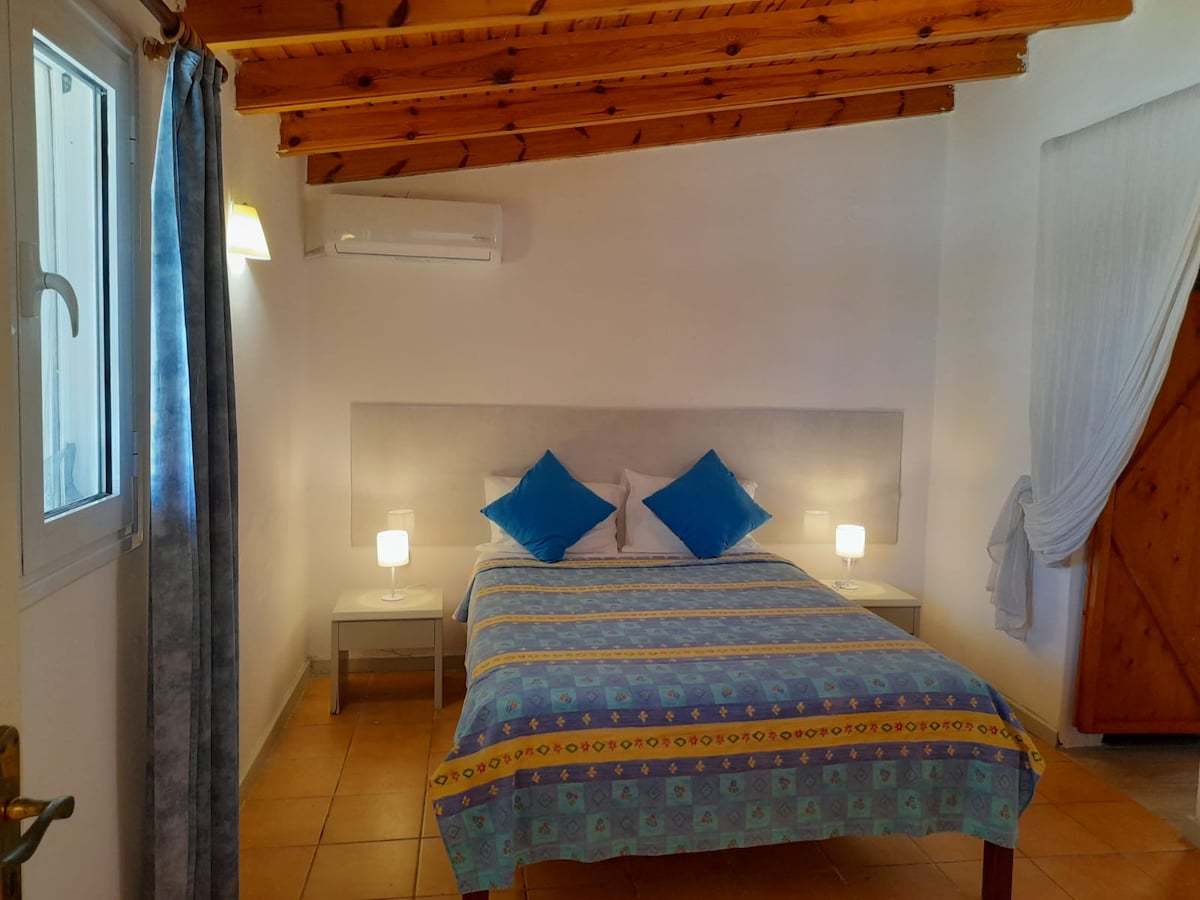 A bright bedroom is displayed with an inviting layout featuring a spacious bed adorned with patterned linens and accent pillows. Soft lighting is provided by lamps on nightstands, and natural light enters through a window dressed with sheer curtains. Wooden beams enhance the ceiling's character.