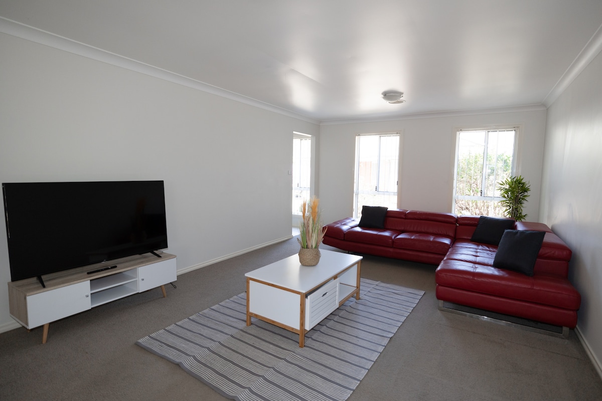 A spacious living area features a large red sectional sofa positioned around a white coffee table. A flat-screen television is set on a media console, while natural light enters through multiple windows, creating a bright and open space.