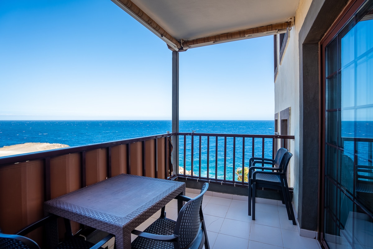 A spacious balcony overlooks the ocean, featuring a table with four chairs. The clear blue sky meets the sea in the distance, creating a serene outdoor space perfect for enjoying meals or relaxation.