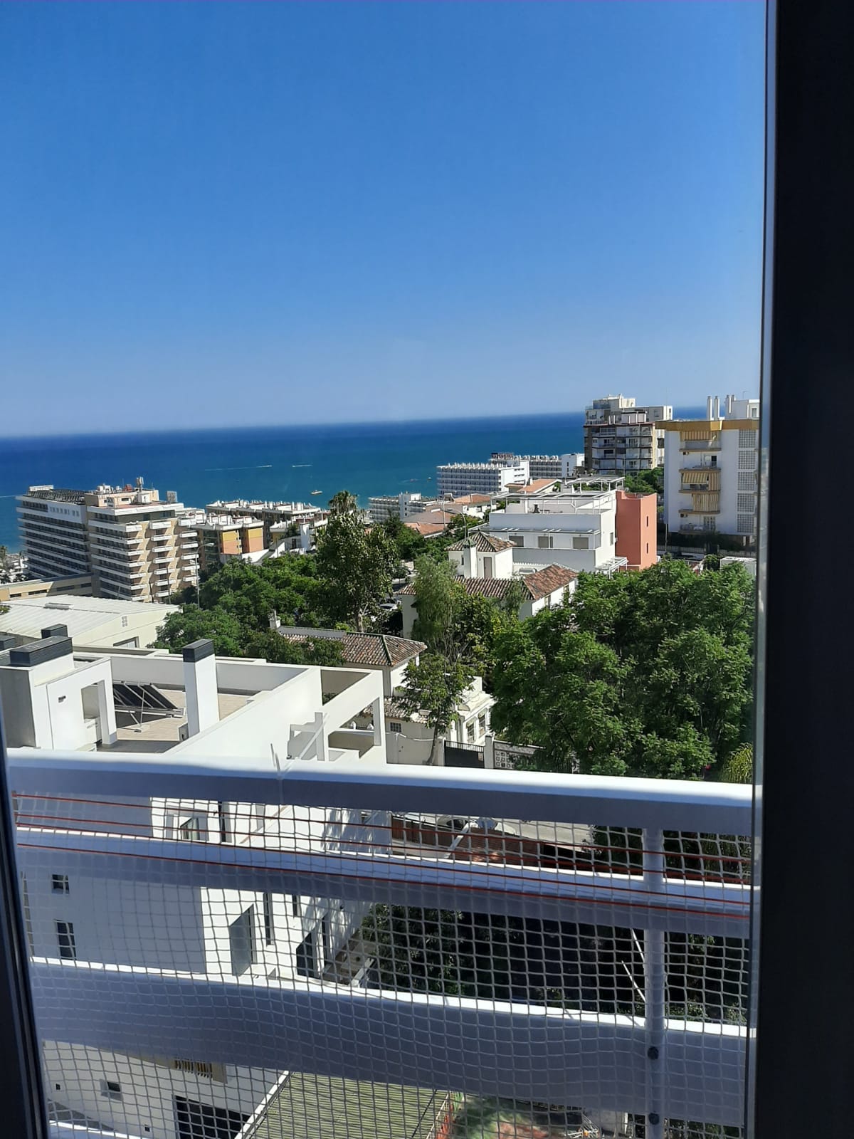 A clear view of the coastline is presented from the balcony, showcasing the blue ocean and several buildings in the foreground. Lush greenery is also visible among the urban landscape, highlighting the blend of nature and city life.