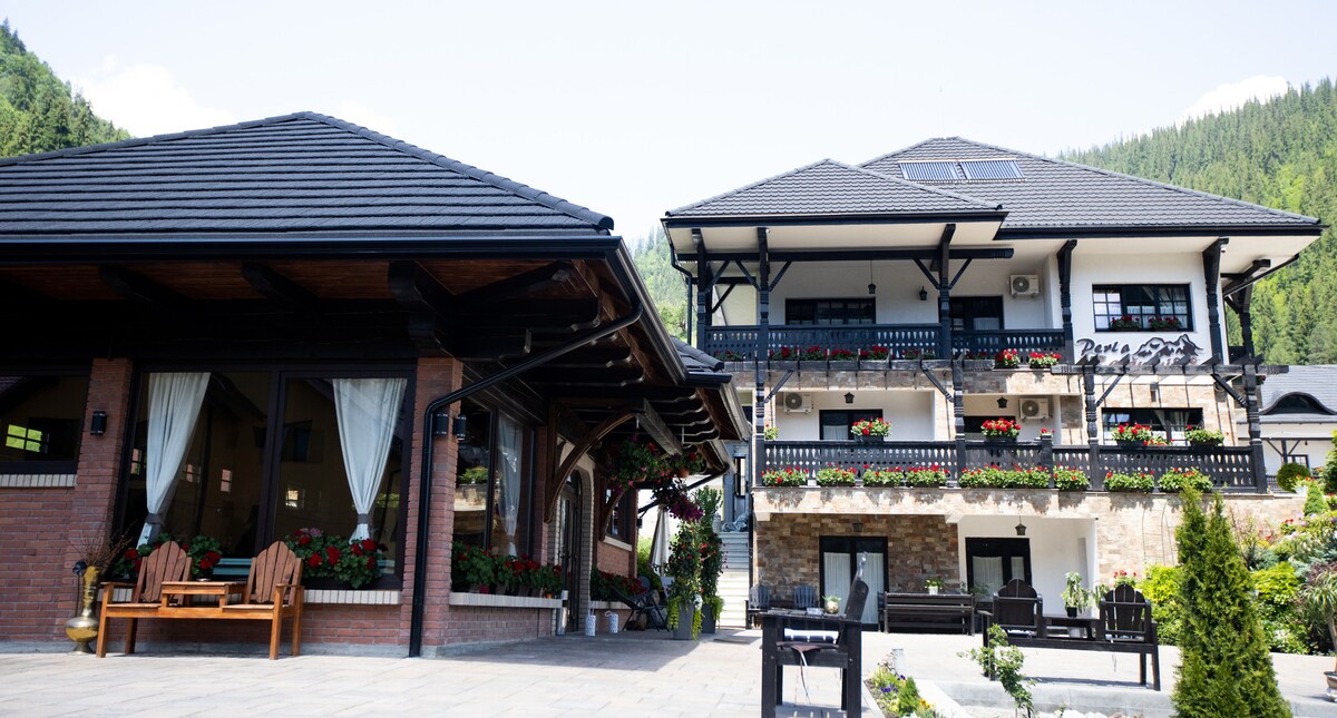 A stylish building featuring a mix of contemporary and rustic architecture, surrounded by lush greenery. The façade includes multiple balconies adorned with vibrant flowers. Spacious outdoor seating areas are available, complemented by sunlit stone pathways.