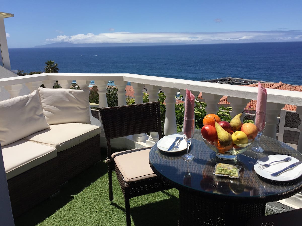 A balcony is depicted with a table set for two, featuring a bowl of fresh fruits. Comfortable seating is available, and the ocean is visible in the background, along with clear blue skies enhancing the serene environment.