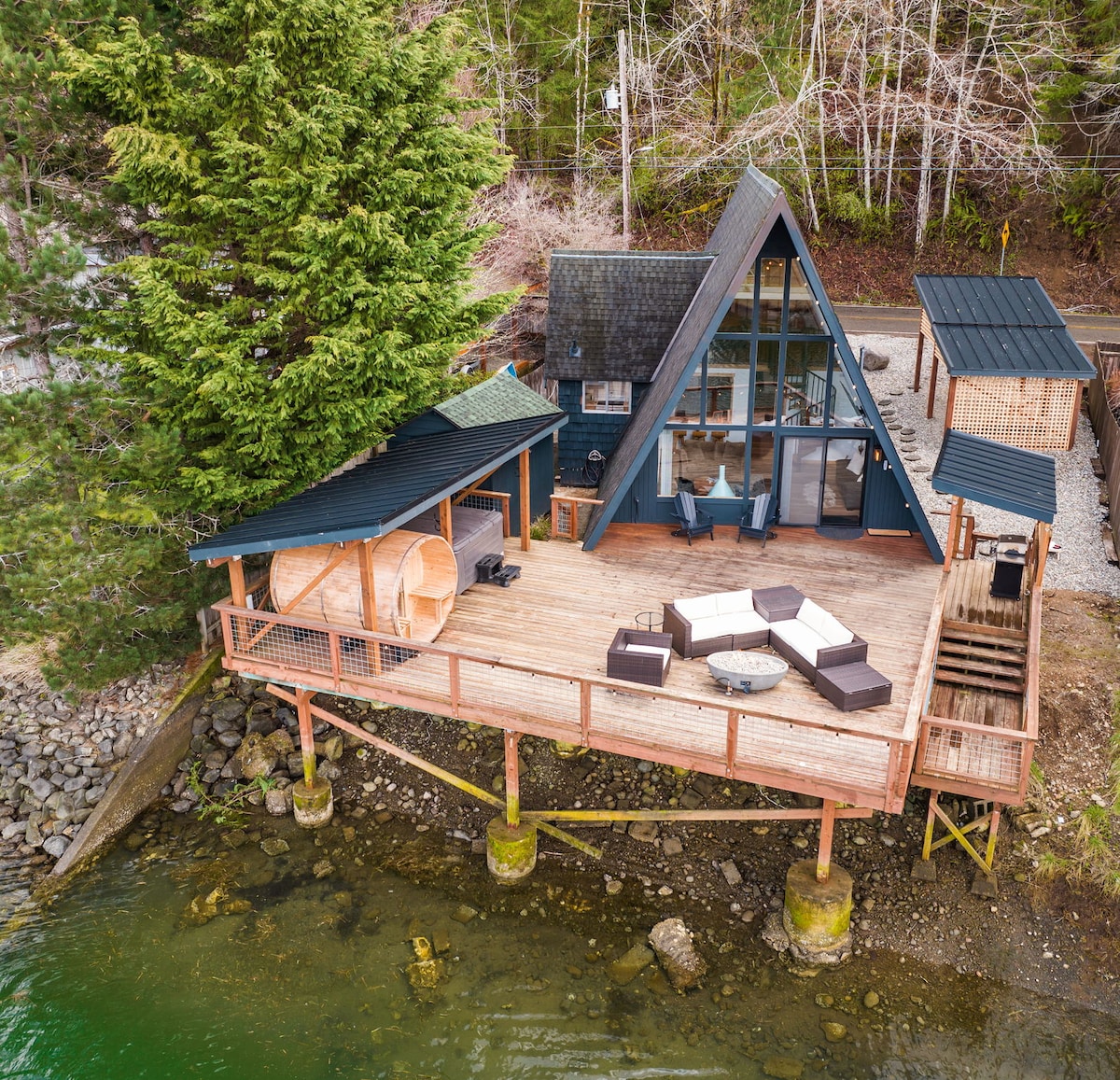 An A-frame home is positioned above the water, featuring a spacious deck with outdoor seating. Nearby, a barrel sauna and steps lead down to the calm water. Surrounded by trees, the property offers a blend of modern architecture and natural beauty.