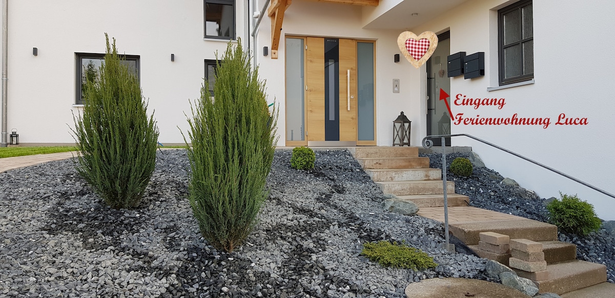 The entrance to Ferienwohnung Luca is depicted, featuring a wooden door framed by sleek glass panels. The pathway is bordered by gravel and ornamental shrubs, leading to a set of stone steps. A mailbox is positioned nearby, contributing to the modern aesthetic.