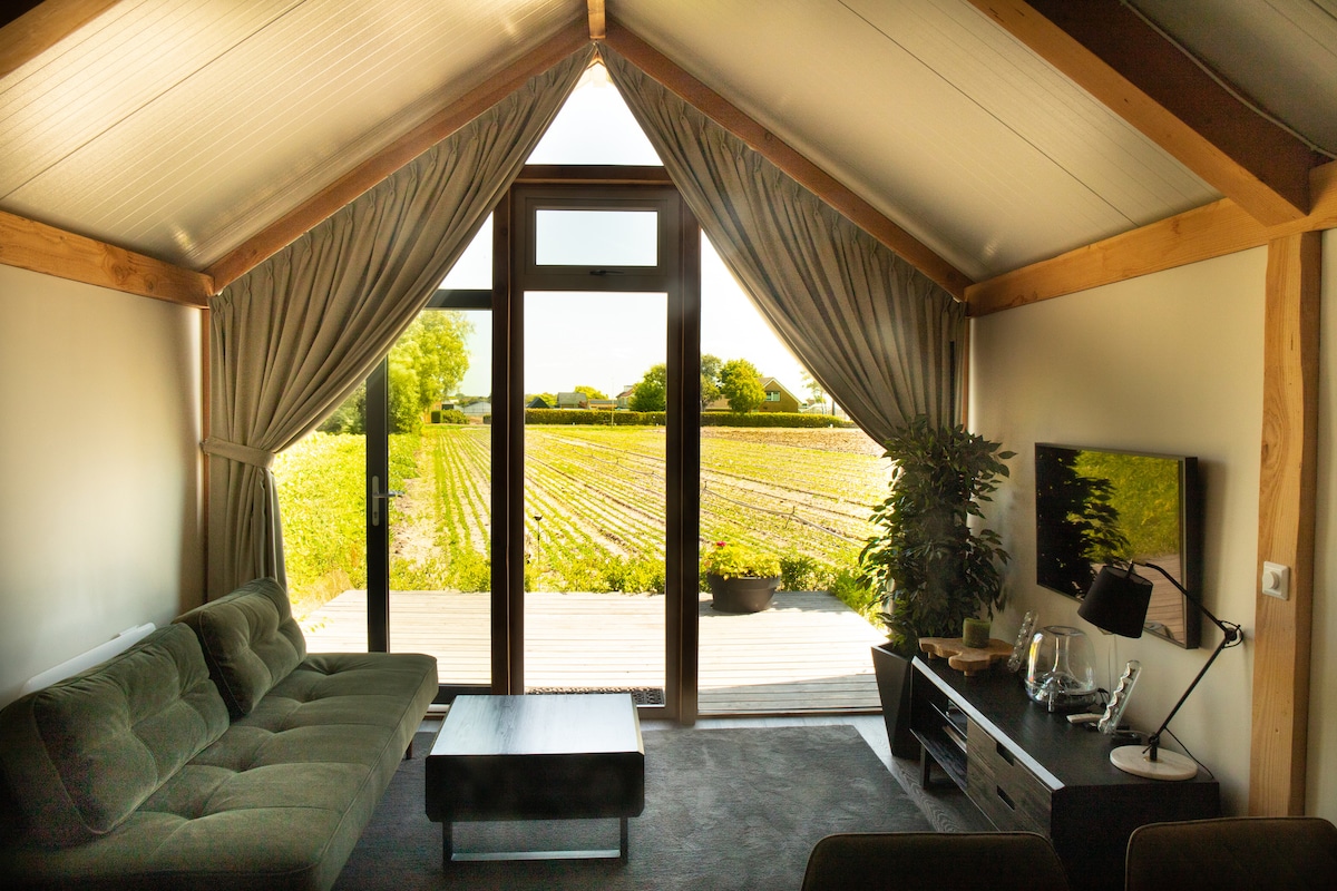 The living area is defined by large glass doors that open to views of the lush fields outside. A comfortable sofa and a coffee table are arranged in front of a media console, creating an inviting space filled with natural light.