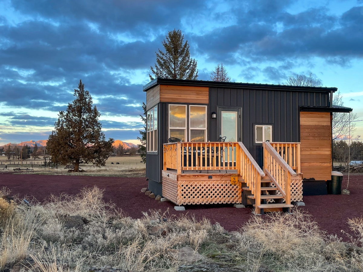 The tiny house exterior is highlighted by wooden siding and large windows, offering views of the surrounding landscape. A welcoming deck with steps leads to the entrance, while the colorful gravel driveway complements the house against a backdrop of dramatic skies and distant mountains.