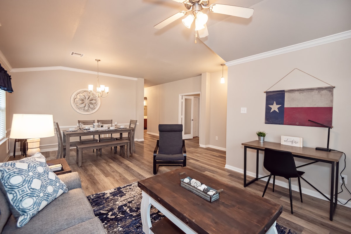 The living space features an inviting layout with a gray sofa and a dark coffee table, complemented by a blue area rug. A dining table is visible in the background, alongside a work desk and a Texas flag decoration. Ceiling fans and natural light enhance the cheerful ambiance.