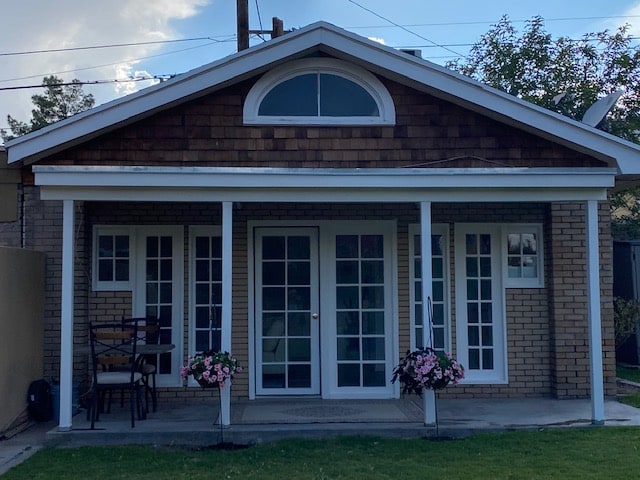 A charming casita is displayed featuring a front porch with two chairs and a small table. The structure showcases large glass doors framed by multiple windows, allowing natural light to illuminate the interior. Pink flowers are arranged in hanging baskets beneath the porch.
