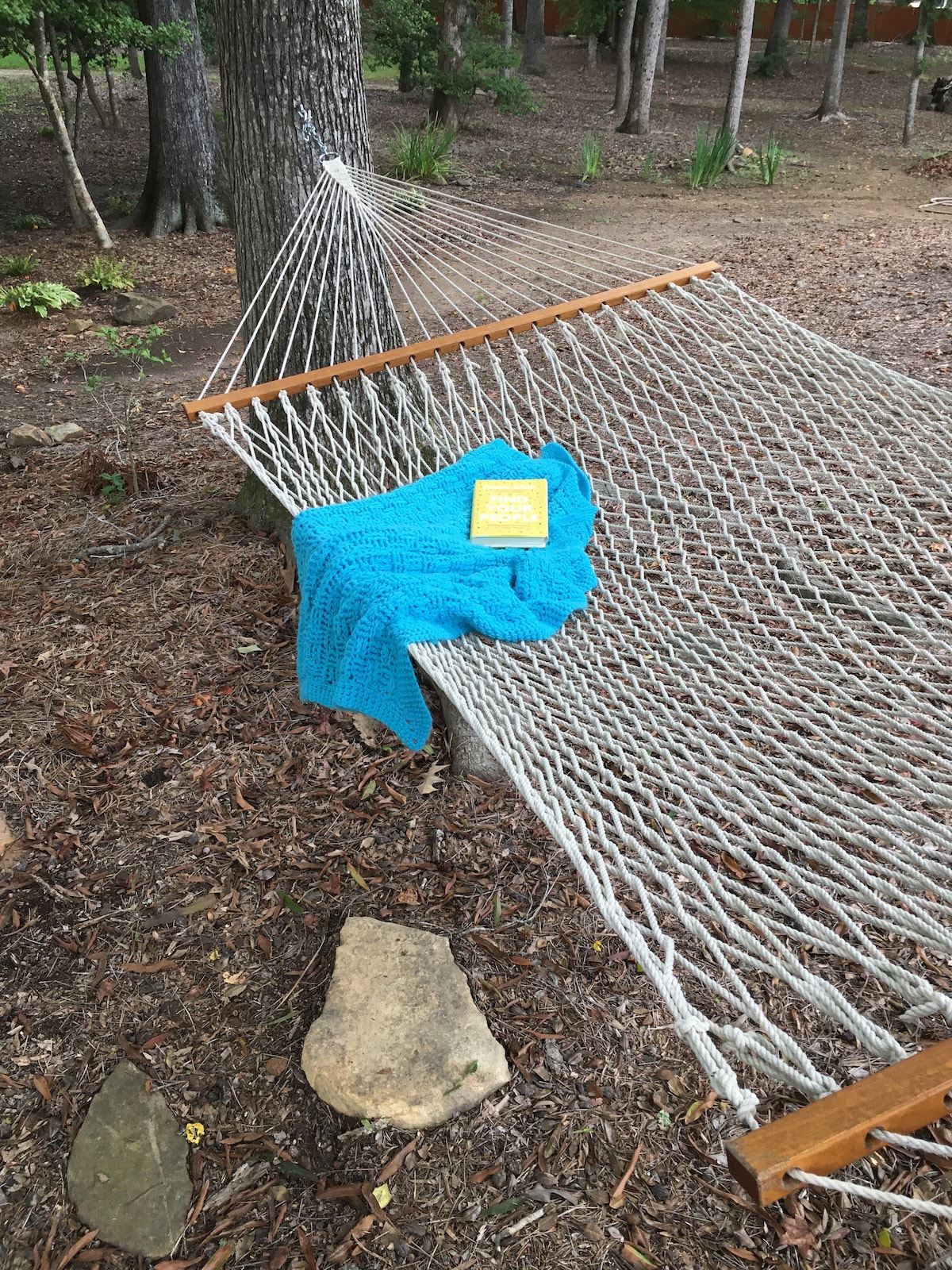 A cozy hammock is nestled between trees, adorned with a bright blue blanket and a book. The surrounding ground is covered with mulch and scattered stones, while greenery creates a serene natural backdrop.