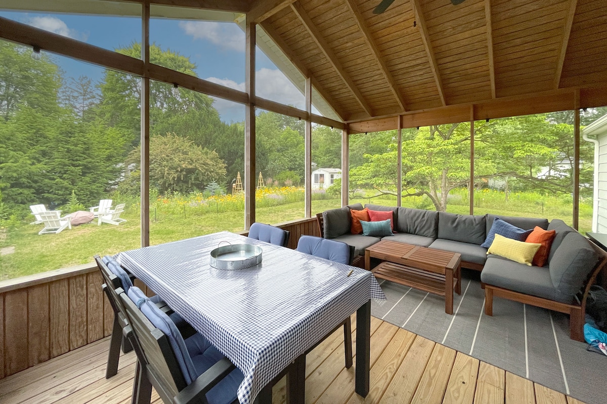 A screened-in porch features a spacious seating area with a sectional sofa adorned in various cushions. A dining table occupies the center, surrounded by a pleasant view of the landscaped yard, including wildflowers and trees in the background.