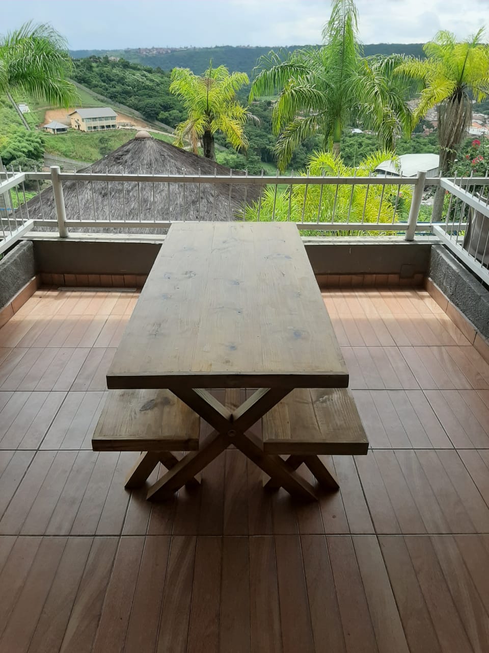 A wooden table and benches are positioned on a spacious balcony, featuring a scenic view of rolling hills and greenery. The terrace is bordered by a white railing, and tropical palm trees are visible in the background, complementing the outdoor dining area.