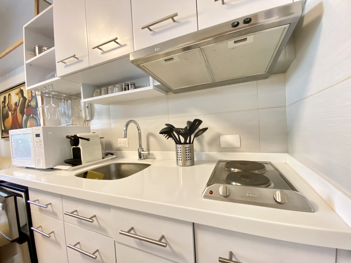 The kitchen features modern white cabinetry and a clean countertop. A stovetop, microwave, and a decorative container for cooking utensils are prominently displayed. Glassware and dishware are neatly arranged on the shelves above.