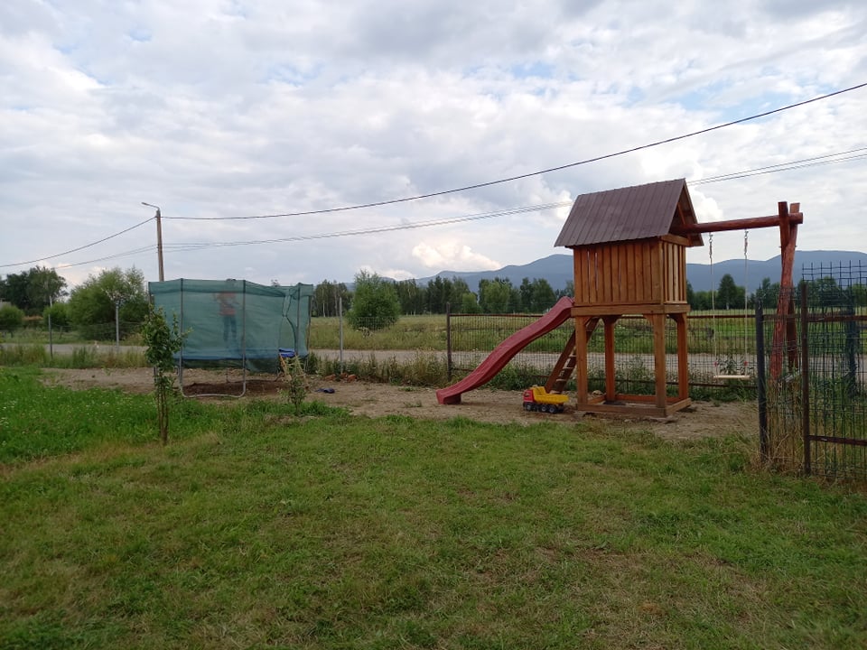 A wooden playhouse stands next to a red slide, positioned on a grassy area. A netted play structure is visible nearby, surrounded by an expansive open space with distant mountains in the background. A boundary fence encloses the area.
