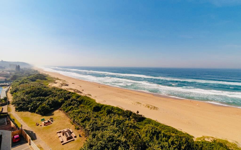 An expansive view of a sandy beach stretches along the coastline, accompanied by rolling waves and a clear blue sky. Lush greenery borders the beach, enhancing the natural scenery, while distant buildings form a subtle backdrop to the serene landscape.