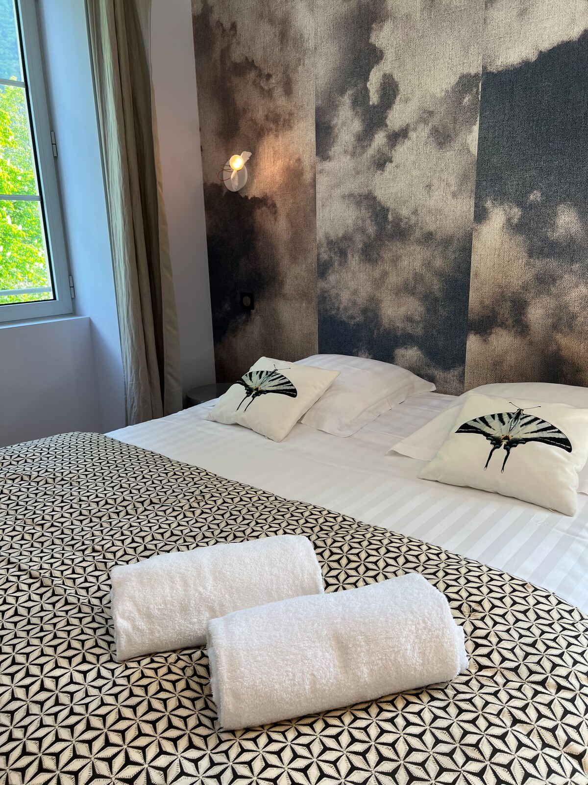 A cozy sleeping area features a spacious bed with a patterned blanket, adorned with decorative pillows. Soft, white towels are neatly arranged on the bed. A large window allows natural light to illuminate the room, highlighting the unique wall design.