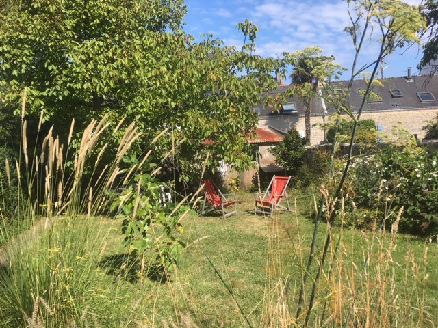A tranquil garden space is portrayed, featuring two red lounge chairs positioned on a lush green lawn. Surrounding greenery, including tall grasses and low shrubs, provides a sense of privacy. A clear blue sky is visible above, enhancing the peaceful atmosphere.