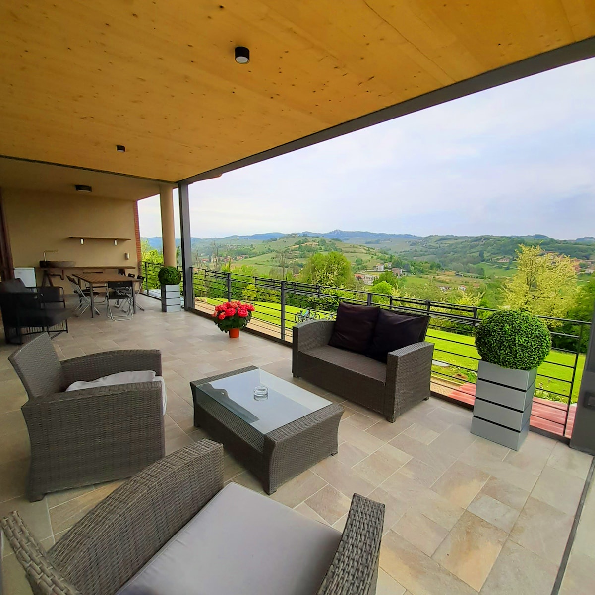 A spacious covered terrace features comfortable seating arrangements, including two armchairs and a central table. Lush greenery and vibrant flowers accent the area, with panoramic views of rolling hills and valleys visible in the background.