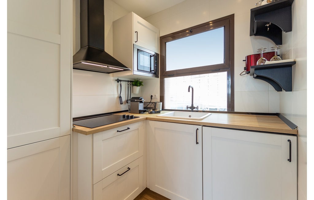 The kitchen features a modern design with white cabinetry and a wooden countertop. A black extractor hood is positioned above the stove, and a window provides natural light. Essential appliances, including a microwave and sink, are neatly arranged to enhance functionality.