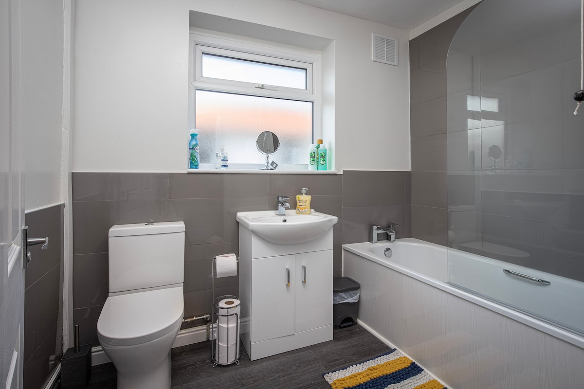 A well-appointed bathroom is showcased, featuring a white toilet and sink set against grey tiled walls. Natural light enters through a window above the sink, while a bathtub is positioned adjacent. Towels and toiletries are neatly arranged on the surfaces.