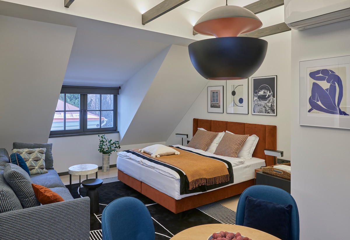 A spacious studio layout is presented, featuring a comfortable sleeping area with a large bed framed by warm textiles. A cozy seating area with modern furniture is visible, and artistic wall decor adds a touch of personality. Natural light filters through a large window.