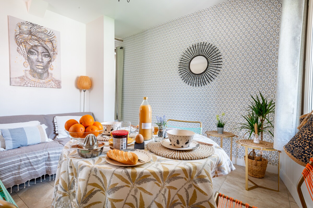 A bright and welcoming living space features a round table adorned with a patterned tablecloth and an array of breakfast items, including bread and fruit. Soft seating is arranged nearby, with decorative pillows adding comfort. A sunlit ambiance is complemented by patterned wallpaper and a stylish wall mirror.