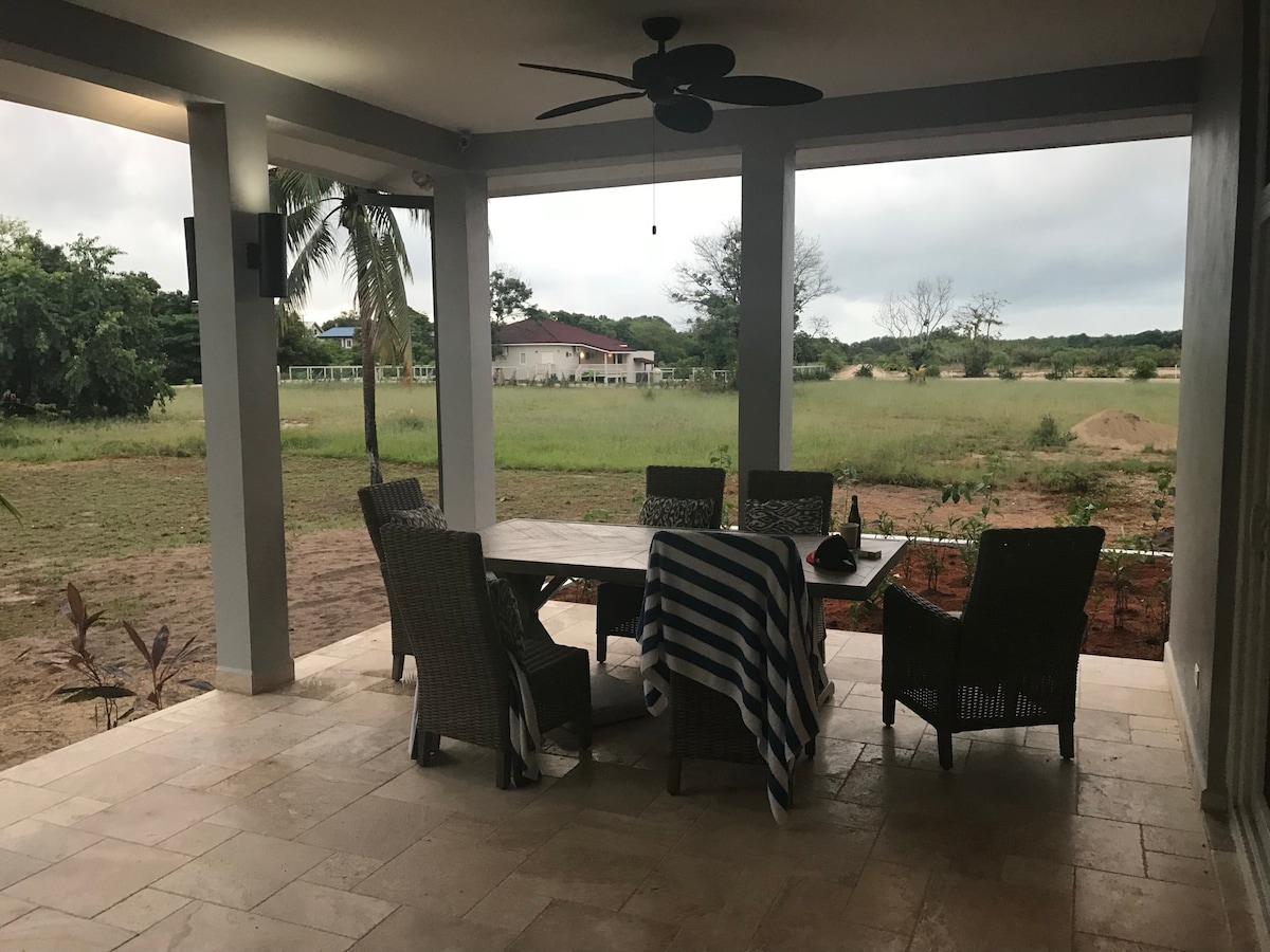 A covered lanai features a dining table surrounded by four wicker chairs. A striped blanket is draped over one chair. The space is open to lush greenery and fields, with a distant view of neighboring homes.