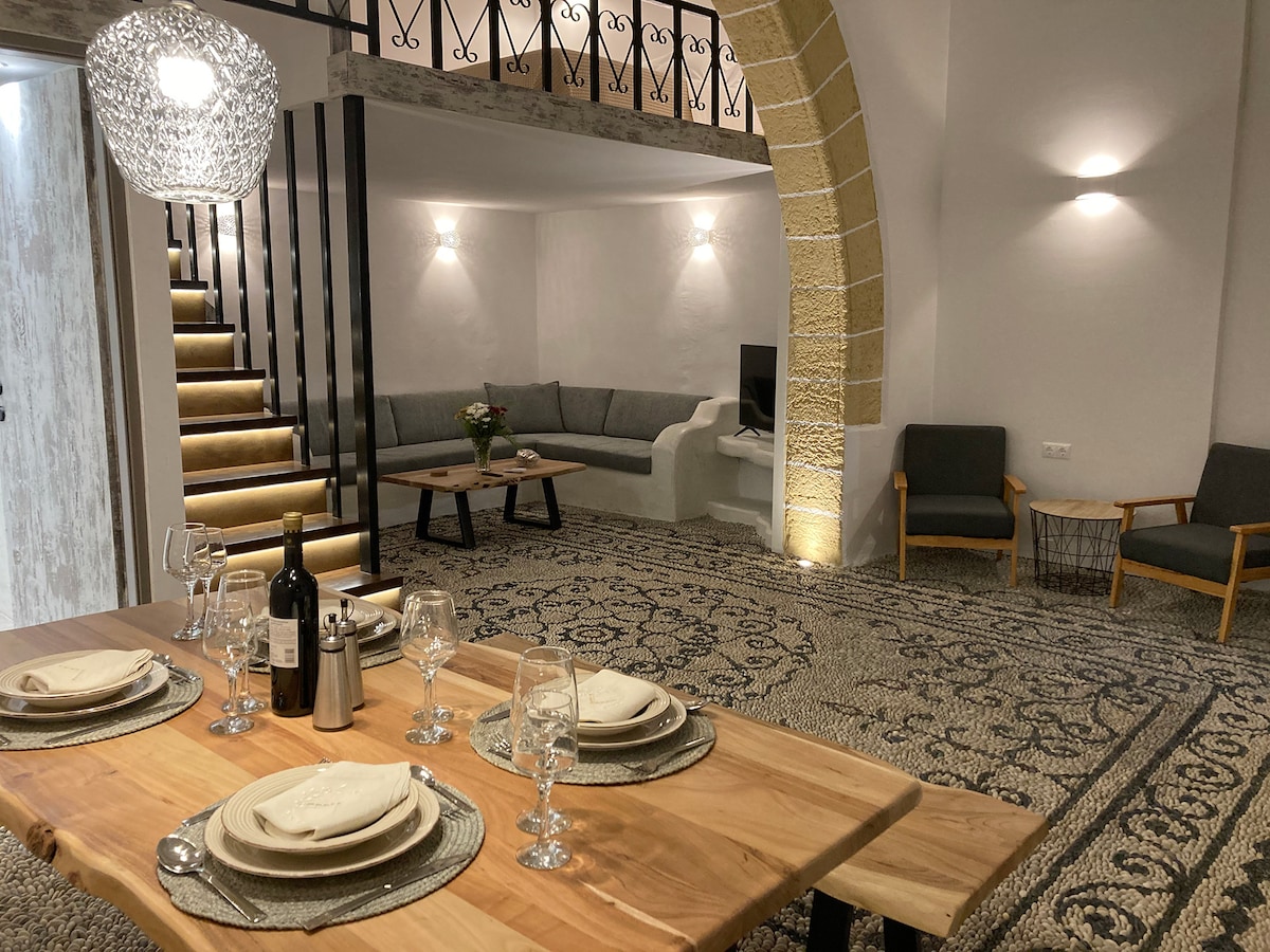 A dining area is presented with a wooden table set for four, featuring glassware and silver utensils. Behind, a comfortable seating area with a gray sofa and decorative cushions is visible. The room is illuminated by soft lighting, highlighting the patterned rug and stone archway.