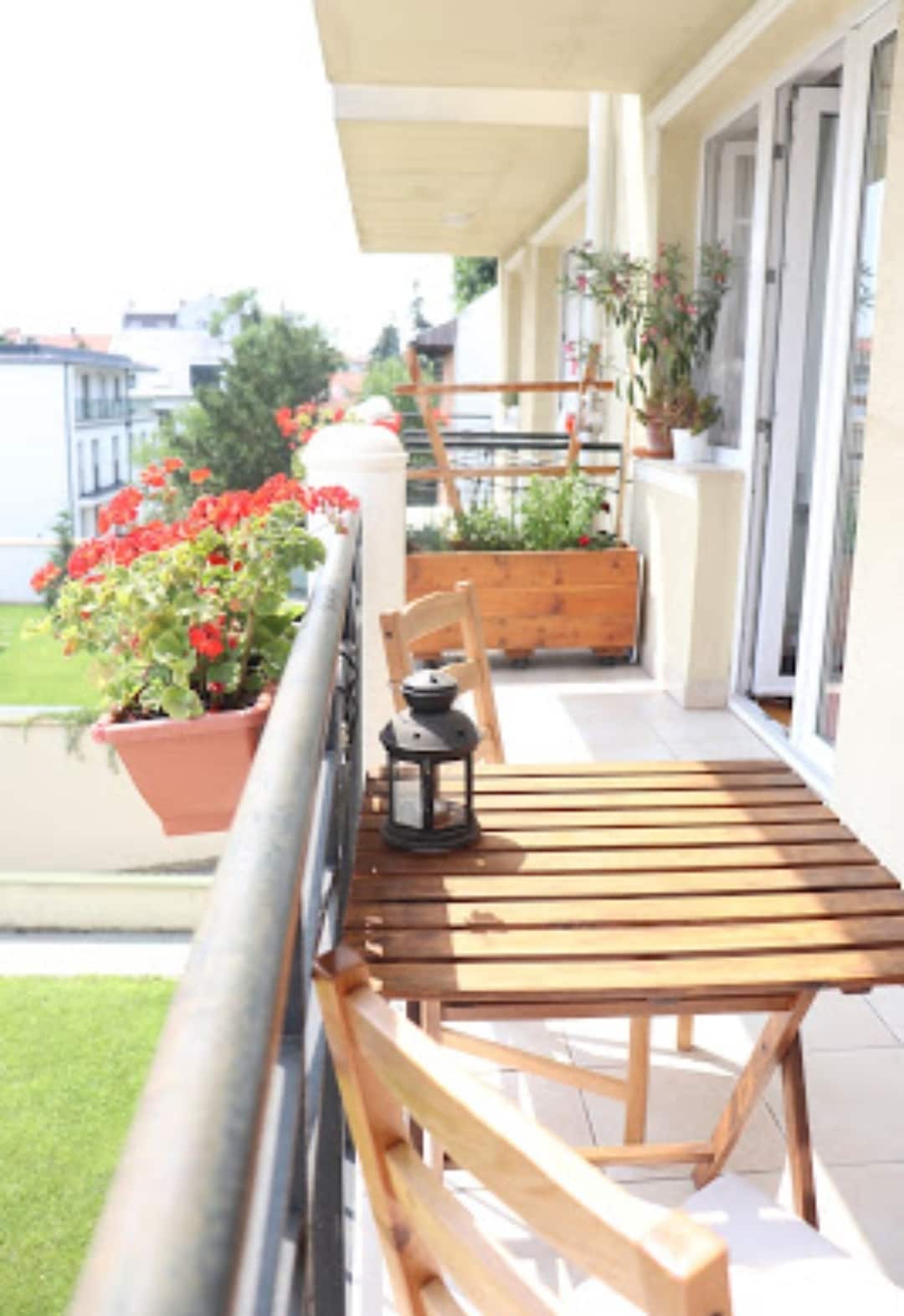 A balcony space is presented with a wooden table and chair, featuring a lantern. Potted plants with vibrant red flowers are seen along the railing, while greenery is visible in the background, providing a serene outdoor setting.
