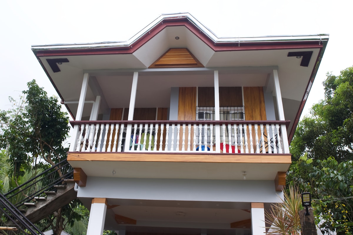 The two-story house features a traditional design with a prominent overhanging roof and a balcony adorned with white railings. Lush greenery surrounds the property, enhancing the natural setting. The entrance is accessible via a staircase leading to the welcoming porch.