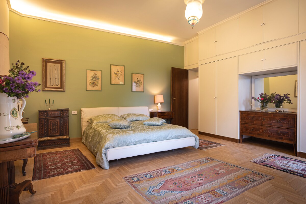 The spacious bedroom is complemented by green walls, featuring a large bed adorned with a light coverlet. Decorative rugs enrich the wooden floor. Natural light enhances the room, while framed artwork and elegant furniture add a refined touch. A closet space is conveniently built-in.