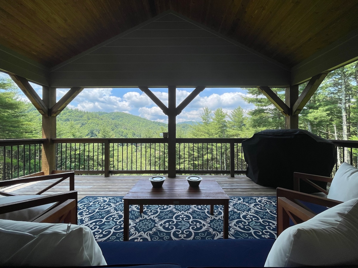 A covered porch features comfortable seating arrangements with a wooden table and plush cushions. A scenic view of the surrounding mountains and trees is captured through the spacious balcony, inviting relaxation in a tranquil outdoor setting.