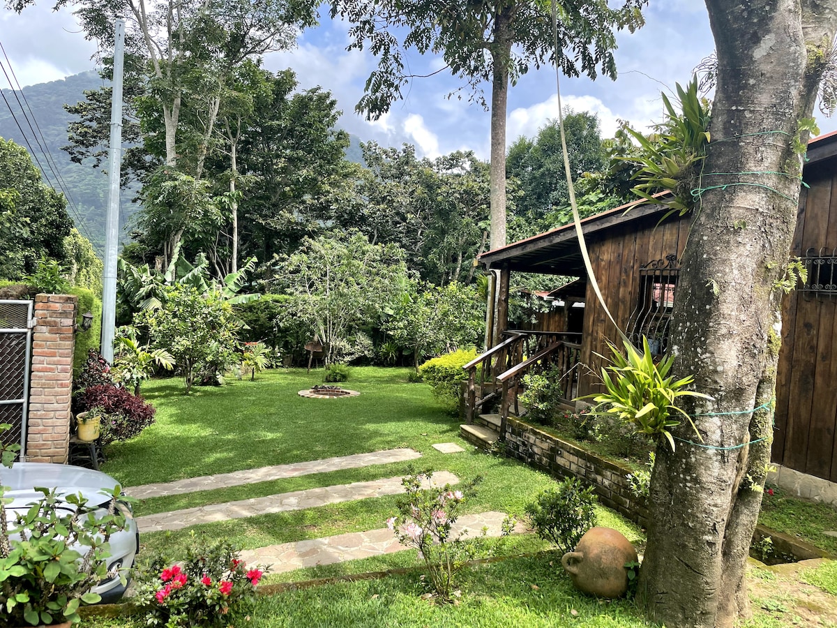 Lush greenery surrounds a rustic wooden cabin, with well-maintained grass and decorative plants enhancing the outdoor area. A stone pathway leads to the cabin, while tall trees provide shade. The serene landscape is complemented by a gentle mountain backdrop.