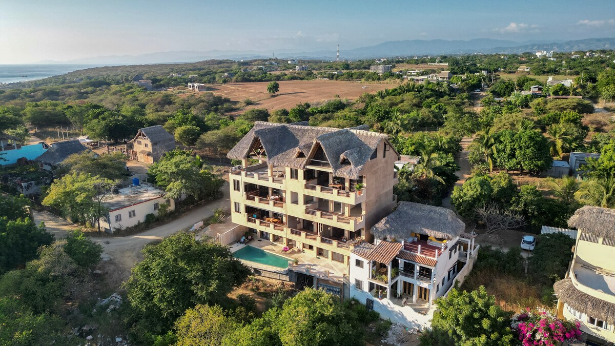 An aerial view showcases the multi-level building nestled within a vibrant landscape. The structure features a thatched roof and multiple balconies, with a swimming pool visible in the foreground. Surrounding greenery and distant hills create a tranquil setting.