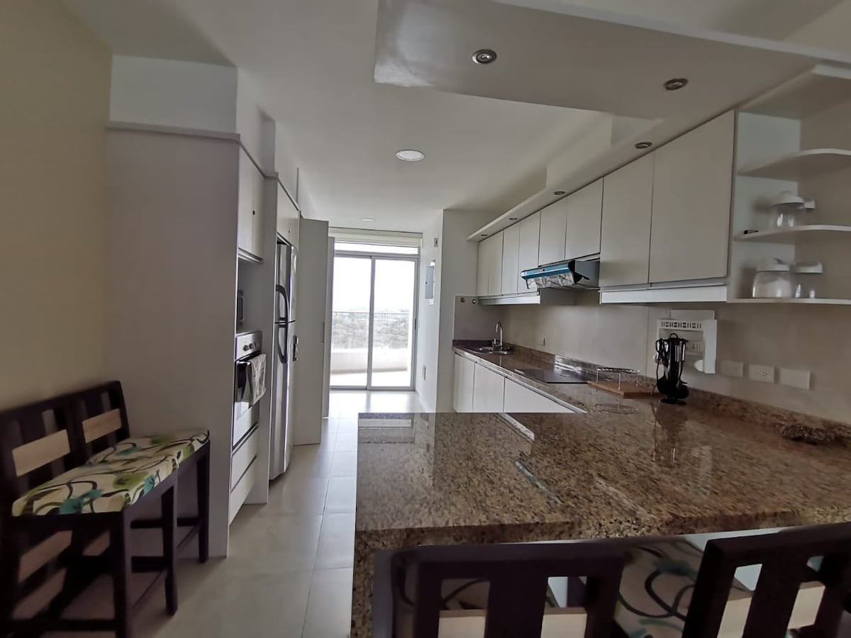A modern kitchen space is displayed, featuring sleek cabinetry and a large granite countertop. Two stools with a patterned fabric are positioned at the counter. A glass sliding door allows natural light to fill the room, providing a view of the outdoor area.