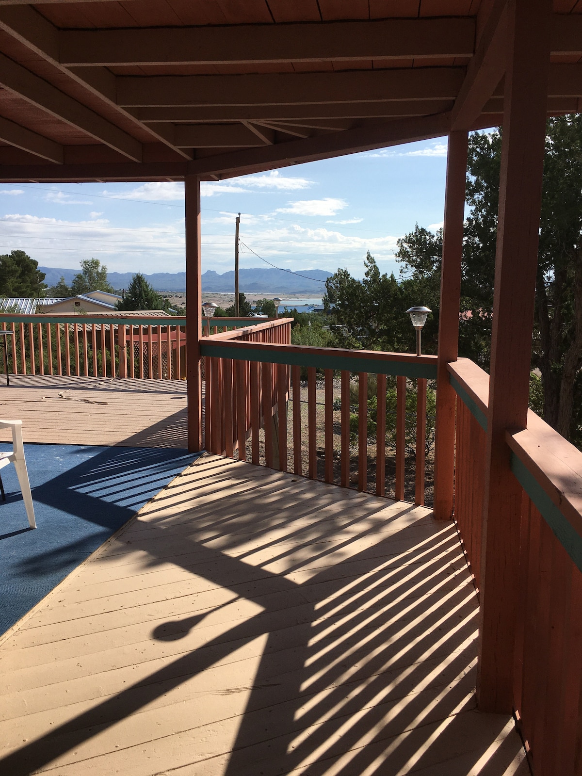 A spacious covered deck with wooden railings is visible, showcasing a view of distant mountains and a clear sky. The deck features a textured floor and is partially shaded by the overhang, creating patterns of light and shadow.