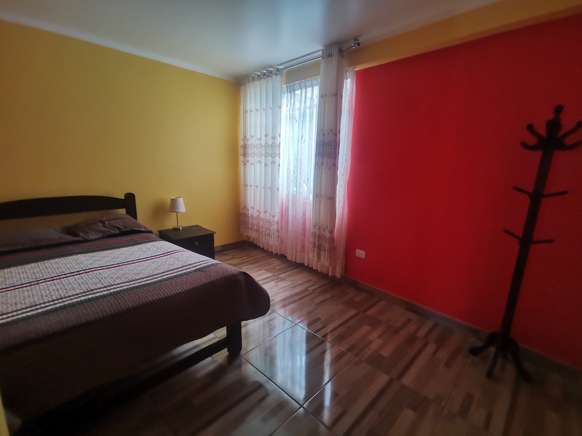 A bright bedroom is designed with warm yellow and red walls. A spacious bed is positioned against the wall, accompanied by a nightstand with a lamp. Natural light filters through sheer curtains, and a coat rack stands in the corner, providing added functionality.