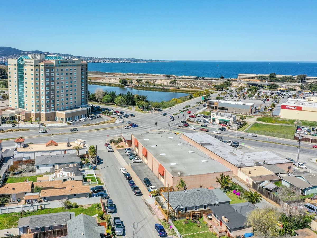 An aerial view captures the area surrounding the coastline. The image showcases a mix of residential and commercial buildings, with a hotel in the background. The Pacific Ocean is visible in the distance, along with a land area that includes a pond and various businesses.