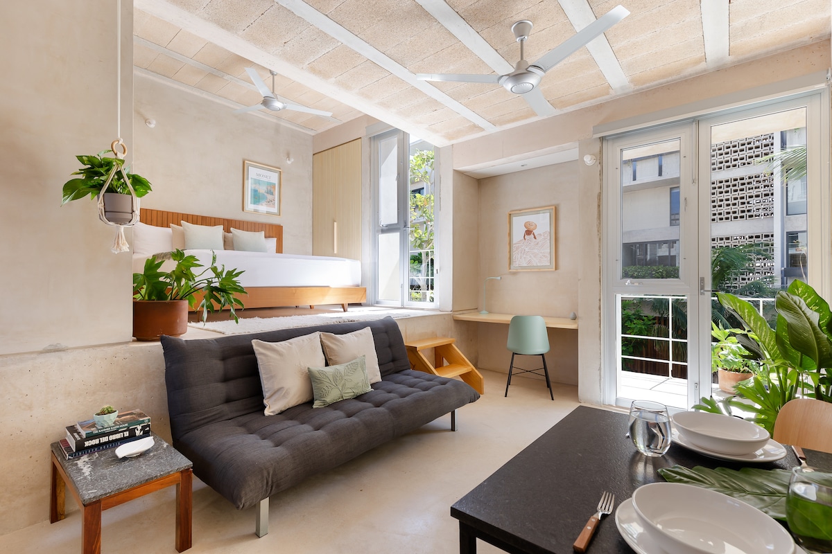 An open living space is featured, showcasing a spacious bed elevated on a platform. A comfortable sofa sits in the foreground, and a small dining area is visible. Natural light fills the room through large windows, enhancing the greenery from outside.