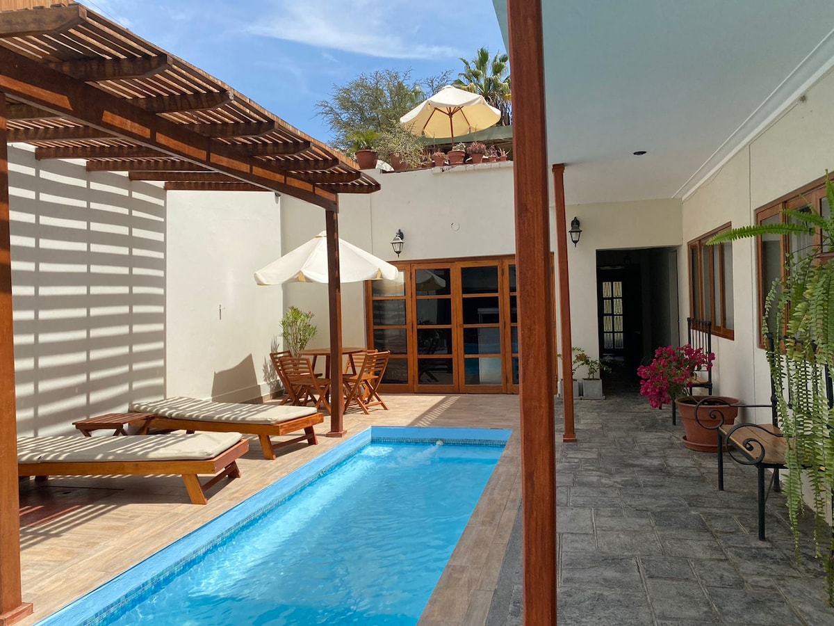 A private pool is surrounded by wooden decking and lounge chairs. Sunlight filters through a pergola, highlighting the inviting blue water. The area includes a small dining set, with potted plants adding touches of greenery. Large glass doors lead into the home.