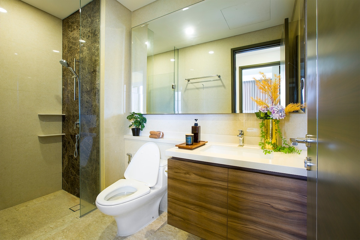 A modern bathroom features a sleek, glass-enclosed shower and a wall-mounted mirror reflecting natural light. The wooden vanity offers storage space, complemented by decorative plants and toiletries arranged neatly. Neutral-toned tiles enhance the clean and sophisticated atmosphere of the space.